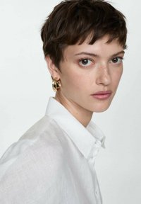 White linen shirt with a pointed collar. Short, dark hair. Gold hoop earring. Natural makeup and neutral background. Brown eyes.
