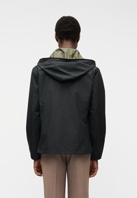 Άνδρας φαίνεται από πίσω φορώντας μαύρο σπορ μπουφάν με κουκούλα πάνω από ένα πράσινο εσωτερικό στρώμα και καφέ παντελόνι, σε λευκό φόντο.