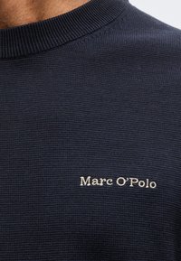 Κοντινό πλάνο σκούτερ πλεκτού μπλε με ριμπ λαιμόκοψη και λευκή κεντημένη επιγραφή "Marc O'Polo" στο στήθος.