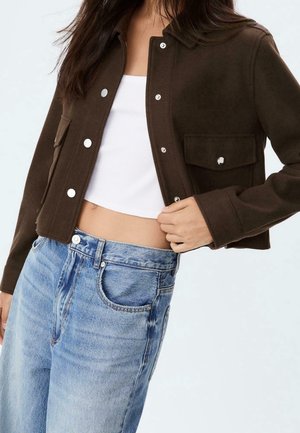 Vrouw draagt een cropped donkerbruine jasje met knopen, een witte cropped top en een lichtblauwe jeans met hoge taille, en houdt het jasje met één hand open.
