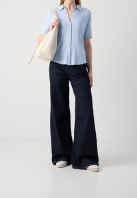 Camicia azzurra a maniche corte con bottoni abbinata a jeans in denim scuri a gamba larga e una borsa a spalla color crema. Sneakers bianche completano il look.