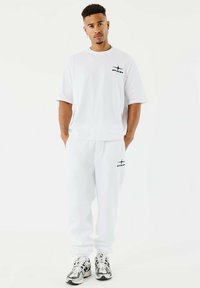 Jonge man met kort, krullend haar die een witte PXP-logo T-shirt en bijpassende joggingbroek draagt, staande met handen in zijn zakken op een witte achtergrond.