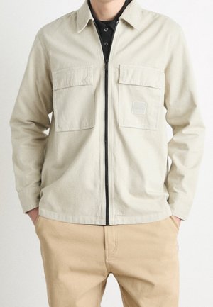 Light beige zip-up jacket with two chest pockets and a logo patch. Made of smooth material with a collar and long sleeves.