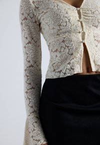 Top corto in pizzo di colore bianco avorio con motivo floreale, maniche lunghe e dettagli in perle. Abbinato a una gonna nera aderente.