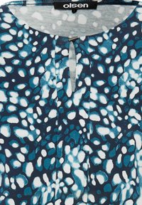 Blaue und weiße gemusterte Bluse mit einem Schlüsselloch-Ausschnitt, aus weichem Stoff und in lockerer Passform. Das Design umfasst unregelmäßige Formen und Akzente.