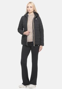 Schwarze Steppjacke mit Kapuze, vorne mit Reißverschluss und gelben Akzenten, getragen über einem beigen Pullover, kombiniert mit schwarzen Hosen.