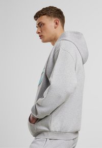 Pelēks dūnu krekls ar lielu tirkīza grafiku, ar auklu kapuci, priekšējo kangaroo kabatu un ribotām piedurknēm un apakšmalu.