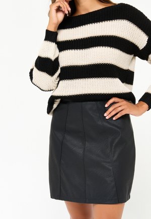 Vrouw die een zwart-wit gestreepte gebreide trui en een zwarte leren minirok draagt, poserend met één hand op de heup en de andere bij het gezicht.