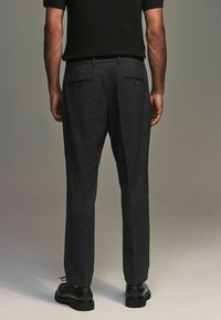 Dark gray trousers made of a textured fabric, featuring two back pockets with button closures and a slim fit design. Black shoes visible.