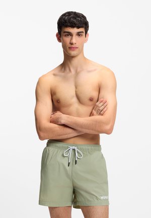 Młody mężczyzna z krótkimi ciemnymi włosami stoi bez koszulki, z założonymi ramionami, ubrany w jasne zielone szorty kąpielowe z sznurkiem i logo "HUGO".