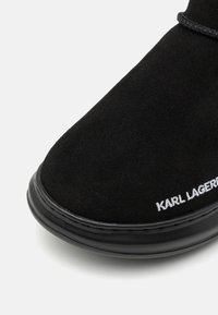 Fekete velúr bokacsizma lekerekített orral, sima fekete talppal és fehér hímzett "KARL LAGERFELD" logóval az oldalon.