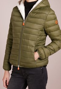 Oliven grøn isoleret jakke med hætte, quiltet design, hvid sherpa-foring, lynlås med orange trækstrop og sidelommer.