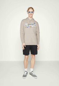Giovane in felpa beige con logo floreale, pantaloni corti neri, sneakers grigie, calzini bianchi e occhiali da sole con lenti azzurre, in piedi su uno sfondo semplice.