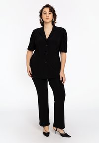 Schwarze Knopfbluse mit kurzen Puffärmeln, kombiniert mit enganliegenden schwarzen Hosen und spitzen schwarzen High Heels mit Knöchelriemen. Schlichtes Design.