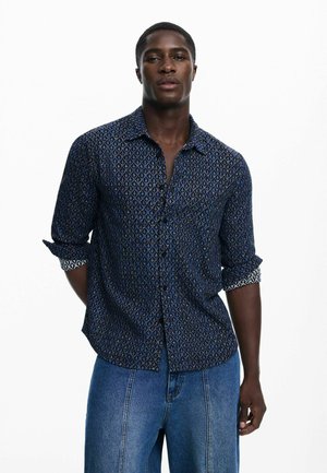 Blå mønstret skjorte med knapper og diamantmønster, med ærmer rullet op og kontrastkant. Båret med lyseblå jeans med lige ben.