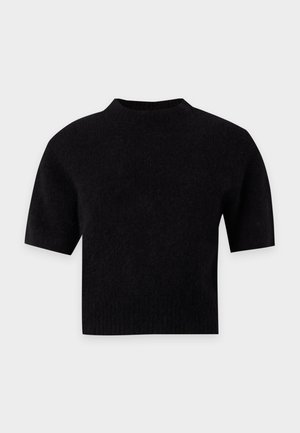 Femme blonde portant un pull noir à manches courtes, un pantalon large taille haute sombre avec ceinture et des chaussures à talons ouverts noires sur fond gris.