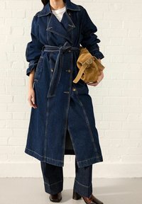 Mujer vistiendo un abrigo de trench de mezclilla oscura y pantalones, con zapatos de cuero marrón, sosteniendo un bolso de cinturón de ante color beige contra una pared de ladrillos blancos.