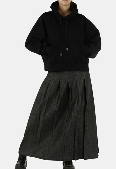 Persoon die een zwarte hoodie en een lange donkere geplooide rok draagt met zwarte laarzen tegen een effen achtergrond.