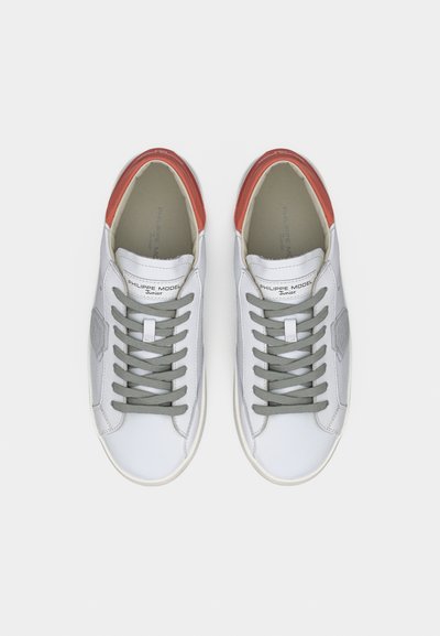 Baskets en cuir blanc avec un accent rouge au talon, des lacets gris et un détail de marque texturé, présentant un design lisse et épuré.