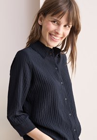 Camicia navy con bottoni, realizzata in un tessuto testurizzato a coste, dotata di colletto a punta e maniche lunghe arrotolate sui polsini. Bottoni neri in vista.
