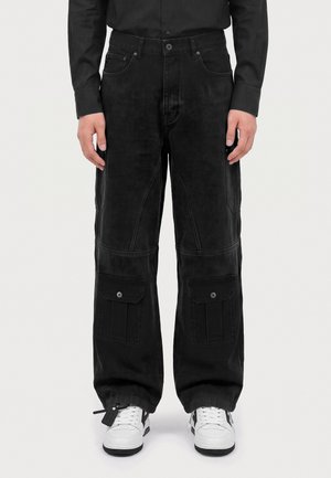 Homme portant un pantalon cargo noir avec des poches à rabat et des baskets blanches, debout devant un fond clair uni.