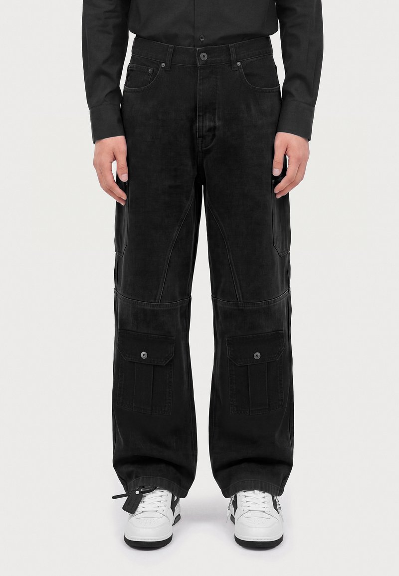 Homme portant un pantalon cargo noir avec des poches à rabat et des baskets blanches, debout devant un fond clair uni.
