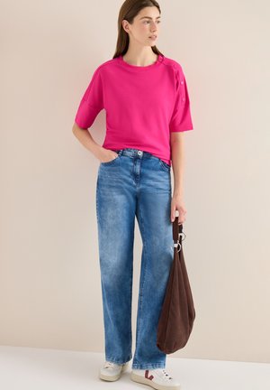 Frau trägt ein knallpinkes kurzärmeliges Oberteil und blaue Jeans, hält eine braune Tasche und steht vor einem einfarbigen beigen Hintergrund.