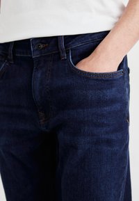 Jean en denim bleu foncé avec une coupe classique, présentant des coutures orange, une poche avant et une fermeture à bouton à la taille.