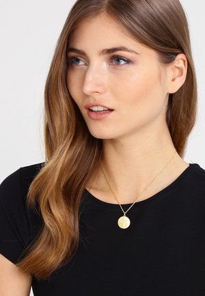 Goldanhänger mit kreisförmigem Design und feiner Gravur, an einer dünnen, zarten Kette getragen, über einem schwarzen Oberteil.