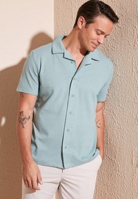 Kortebloezige, lichtblauwe, gestructureerde knopshirt met een afgeknipte kraag en contrasterende donkere accenten. Graag gedragen met witte broeken.