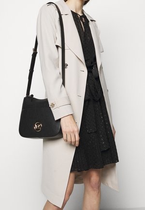 Vrouw draagt beige trenchcoat over een zwarte jurk met patroon, en draagt een zwarte leren schoudertas met gouden ronde sluiting.