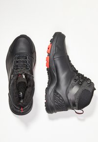 Mustat vaelluskengät, joissa on sileä nahkapinta, teksturoituja kangaspaneeleja, kumipohjat punaisilla yksityiskohdilla ja vahvistetut nauha-aukot.