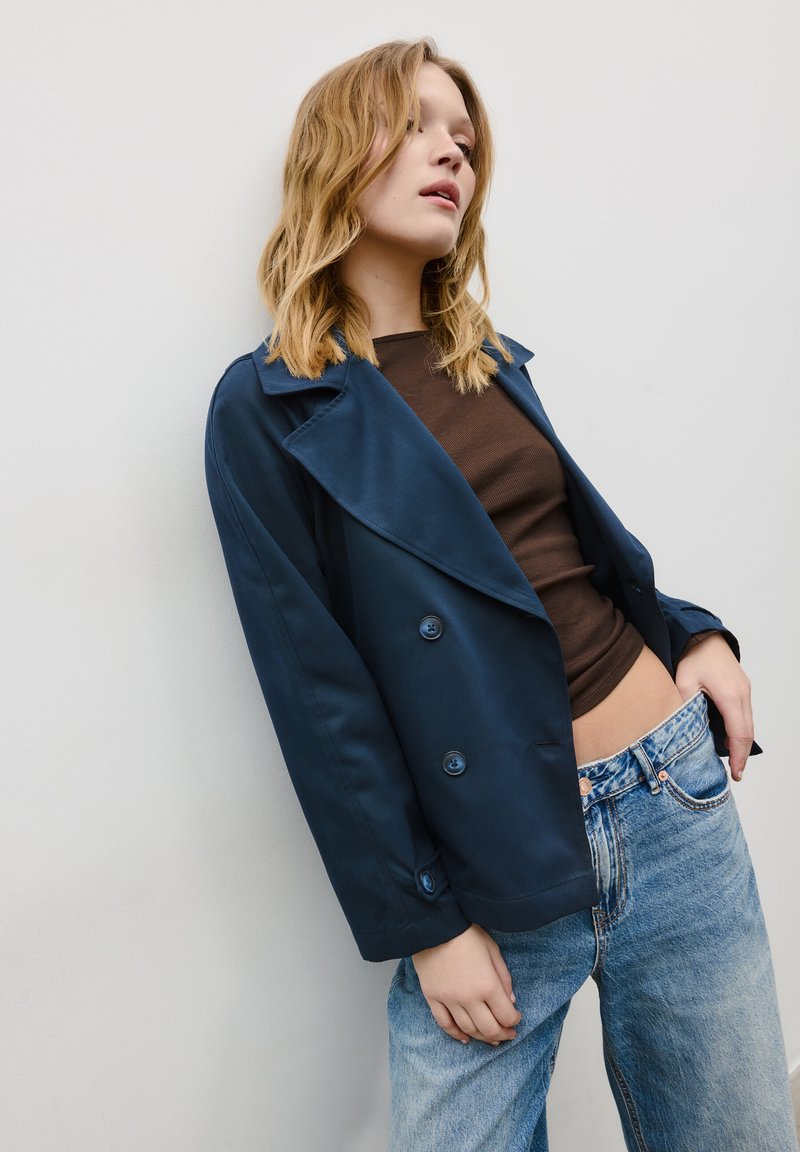 Jeune femme aux cheveux blonds portant un manteau bleu marine, un haut marron et un jean bleu, appuyée contre un mur blanc, la main dans la poche.