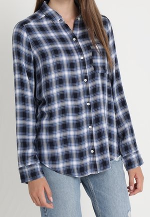 Vrouw draagt een blauw-zwart ruitjes overhemd met knopen en lange mouwen, gecombineerd met lichtblauwe gerepareerde jeans, tegen een effen achtergrond.