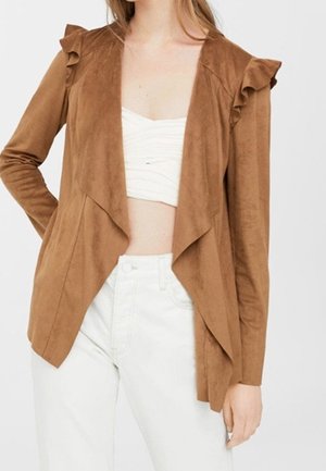 Vrouw draagt een bruine suède jas met open voorkant en ruches op de schouders, een wit gerimpeld crop top en een witte broek met hoge taille.