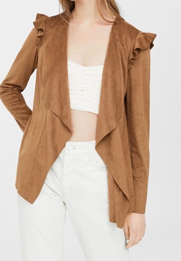 Femme portant une veste en suède marron ouverte à l'avant avec des détails à volants sur les épaules, un crop top blanc froncé et un pantalon blanc taille haute.