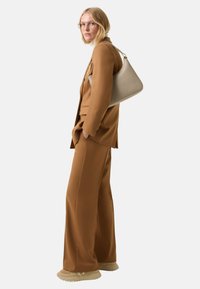 Blondyna w okularach, ubrana w brązowy garnitur, trzyma beżową torbę na ramię, stoi na tle białego tła.