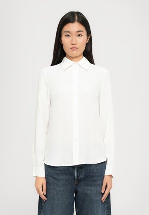 Hvit langermet bluse med krage, laget av et lett stoff. Har knapper foran og mansjetter med rett kant.