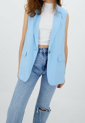 Vrouw draagt een mouwloos lichtblauw blazer over een witte geribde crop top en lichtblauwe high-waist jeans met een scheur op de knie, staand tegen een effen achtergrond.