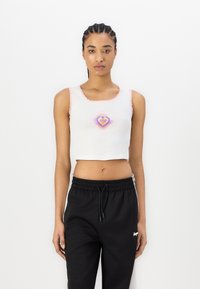 Hvit croppet tanktopp med rosa bølget kant, med et hjerte-design i lilla og gult. Kombinert med sorte joggebukser med snøring.