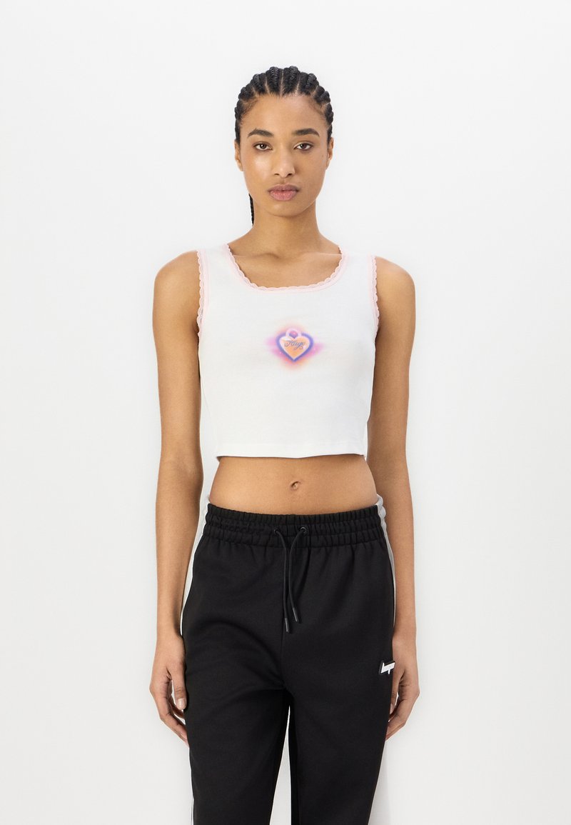 Hvit croppet tanktopp med rosa bølget kant, med et hjerte-design i lilla og gult. Kombinert med sorte joggebukser med snøring.