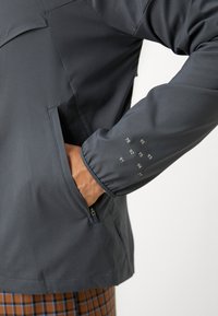 Personne portant une veste gris foncé avec la main dans la poche latérale zippée, montrant une manche imprimée de petits chiffres blancs.