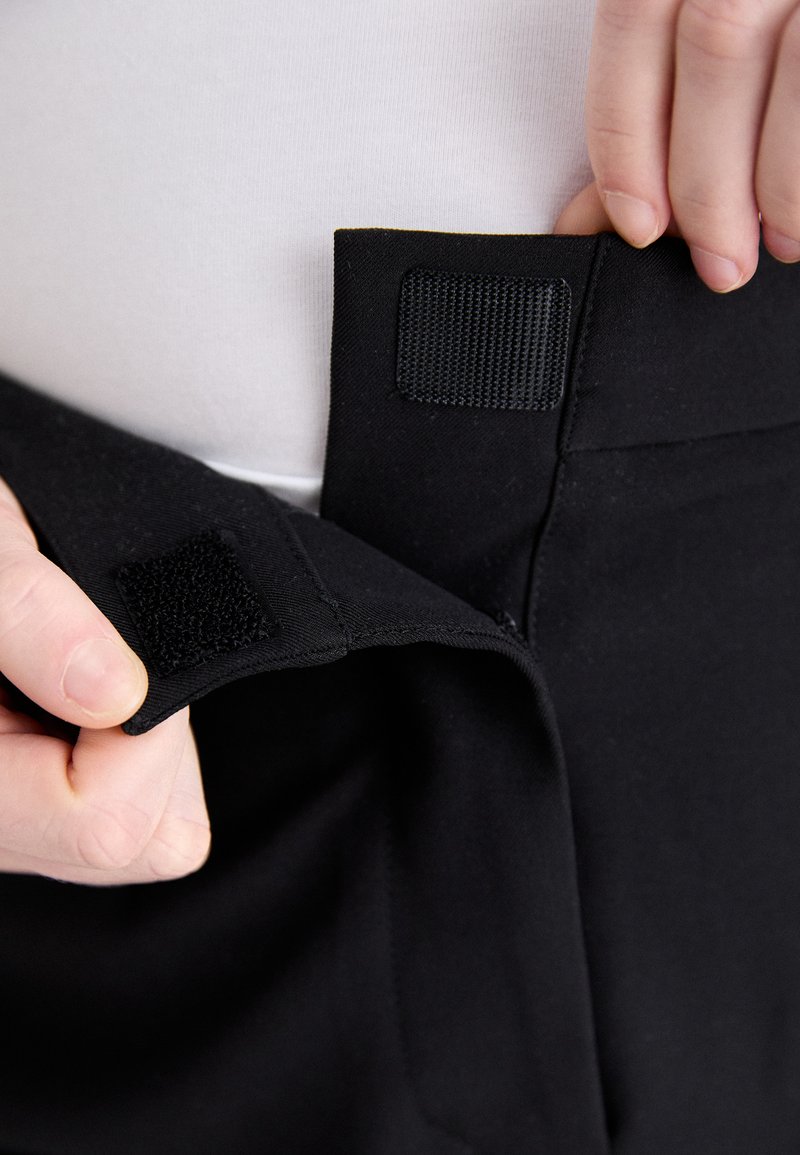 Čierne nohavice sa zapínajú na suchý zips v páse. Tkanina je hladká a elastická, zobrazená proti bielej košeli. Detailný záber na zapínanie.