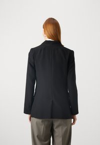 Musta räätälöity blazer, jossa on rakenteellinen muotoilu, uurrettu kaulus ja yksinkertainen halkio takana, yhdistettynä harmaisiin leveisiin housuihin.