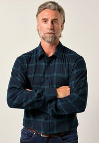 Mörkblå flanellskjorta med teal-rutigt mönster, knäppt, långärmad, och bröstficka. Bärs med blå jeans och brunt bälte.