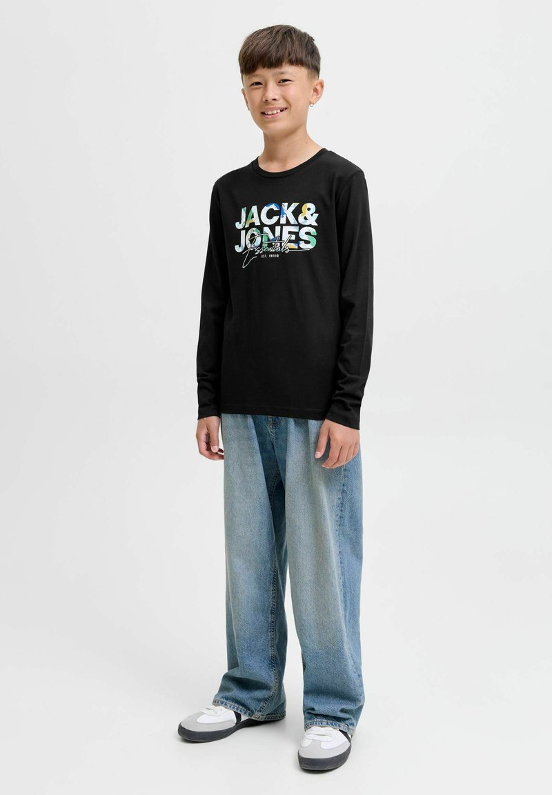 Fiú áll, és mosolyog, fekete hosszú ujjú pólót visel, rajta "Jack & Jones" felirattal, világoskék, bő farmert és fehér sportcipőt.