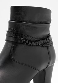 Bota negra hasta el tobillo en cuero sintético liso con punta punteada, tacón alto y correa decorativa texturizada con detalles en negro.