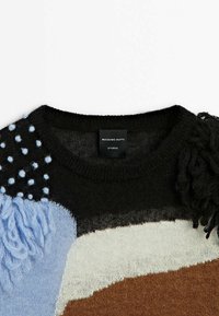 Strickpullover mit einer Farbzusammenstellung aus Schwarz, Hellblau, Beige und Braun. Texturierte Akzente umfassen flauschige blaue und schwarze Fransen sowie Bommeln.