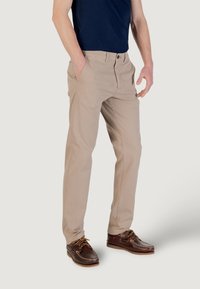 Pantaloni di cotone beige con tasche laterali, chiusura a bottone e passanti per cintura, abbinati a scarpe marroni. Design slim-fit, texture liscia.
