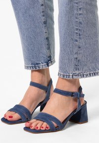 Blaue Wildleder-Absatzsandalen mit zwei breiten Riemen �über den Zehen, Knöchelriemen und einem quadratischen Blockabsatz, getragen mit einer verkürzten Jeans.